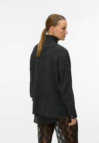 Tmavě šedý svetr s vysokým límcem, volným střihem a žebrovanou texturou. Disponuje dlouhými rukávy a designem se spadlými rameny, nošený přes černé krajkové kalhoty.