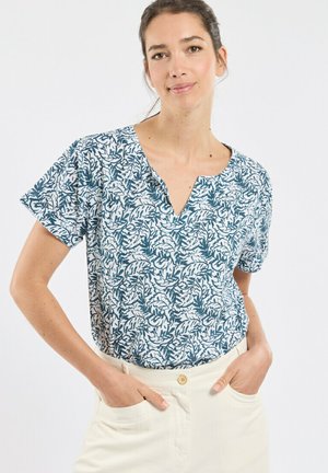 Femme aux cheveux foncés portant un chemisier à manches courtes bleu et blanc à motifs, un pantalon clair, les mains dans les poches, debout devant un fond blanc.