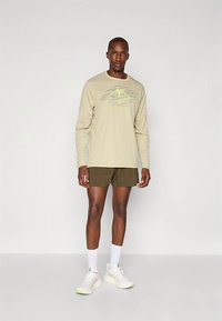 Beige shirt met lange mouwen en een groene bergafbeelding, gecombineerd met olijfgroene korte broek. Model draagt witte sokken en lichtgekleurde sneakers.