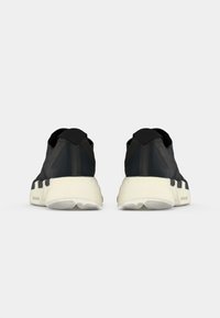 Et par sorte sneakers med hvide såler set bagfra, der viser et minimalistisk design og en tyk dæmpet sål med branding på siden.