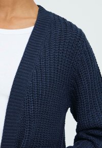 Marineblå strikket cardigan med et struktureret mønster, åben front og ribbet kant. Stoffet virker blødt og tykt, velegnet til lagdeling.