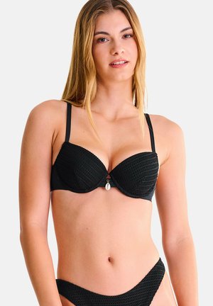 Jeune femme aux longs cheveux blonds portant un bikini noir texturé avec un petit pendentif en coquillage au centre du haut du bikini.