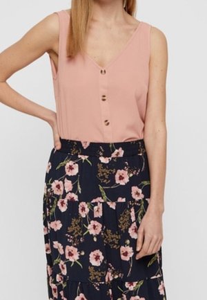 Frau trägt ärmellose rosa Bluse mit Knöpfen und marineblauen Rock mit rosa Blumenmuster, Hand auf der Hüfte ruhend.