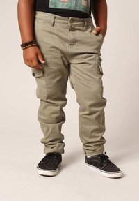 Khaki cargobroek met een knoop- en ritsluiting, meerdere zijzakken en een relaxte fit, gecombineerd met zwarte sneakers.