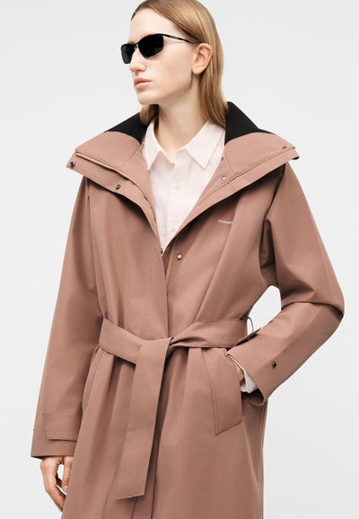 Žena v pripnutom prachovozelenom kabáte so širokým golierom, bielou košeľou a čiernymi slnečnými okuliarmi, ktorá sa pozerá do strany.