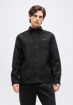 Νέος άνδρας που φοράει μαύρο πουκάμισο με κουμπιά, με υφασμένη τσέπη στο στήθος και το λογότυπο "BOSS" στην αριστερή πλευρά, στέκεται μπροστά σε ένα απλό φόντο.