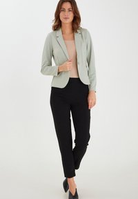 Světle zelený dobře padnoucí blazer s jednoknutíkovým zapínáním, spárovaný s černými kouskovanými kalhotami. Jednoduchý design, hladká textura a žádné viditelné vzory.