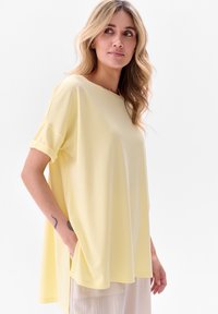 Żółta, oversize bluzka z krótkim rękawem, wykonana z miękkiego materiału, z podwiniętymi mankietami i boczną kieszenią. Gładka faktura i luźny krój.