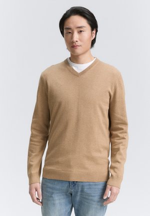 Suéter de punto color marrón claro en forma de V, con mangas largas y dobladillo acanalado. Llevado sobre una camisa blanca, con un sutil detalle de logotipo.