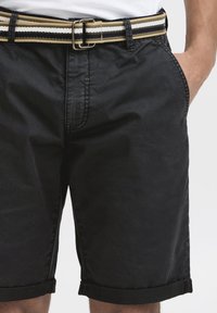 Black denim shorts with folded cuffs, paired with a multi-striped woven belt in olive, white, and black, featuring a silver metal buckle.