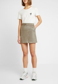 Khaki semišová mini sukně s kapsami, spárovaná s bílým bavlněným tričkem s černým grafickým designem. Boty jsou bílé s barevnými akcenty.