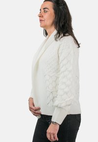 Mujer con cabello oscuro vestida con un cárdigan blanco de punto texturizado y pantalones negros, de pie de perfil contra un fondo blanco.
