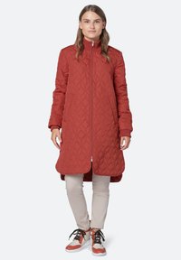 Abrigo largo acolchado rojo con cuello alto y cierre de cremallera. Presenta bolsillos laterales y puños acanalados. Lleva puesto sobre pantalones beige y zapatillas coloridas.