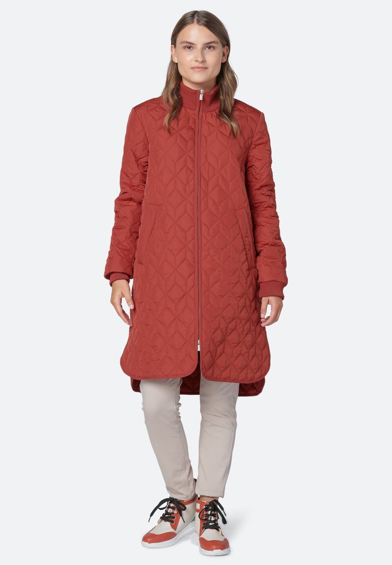 Abrigo largo acolchado rojo con cuello alto y cierre de cremallera. Presenta bolsillos laterales y puños acanalados. Lleva puesto sobre pantalones beige y zapatillas coloridas.