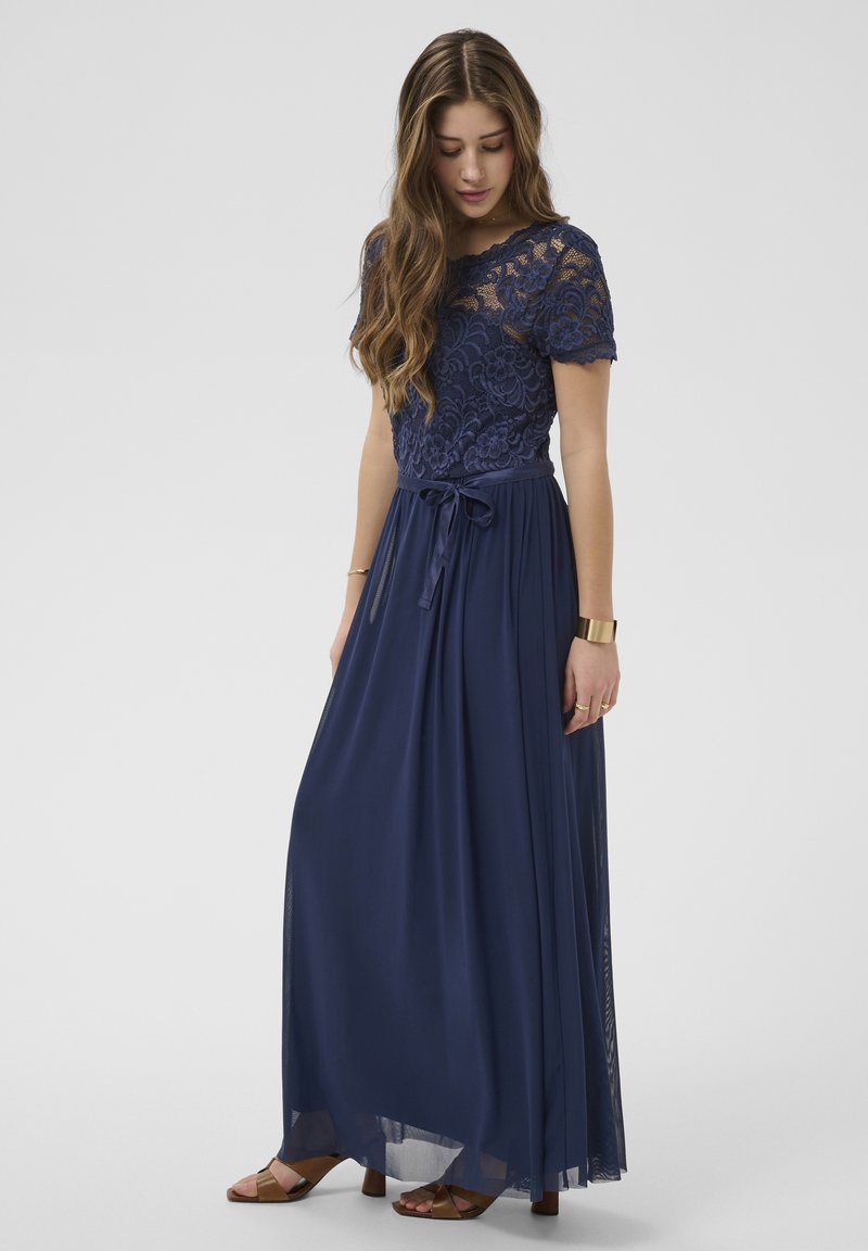 Jeune femme portant une robe en dentelle bleu marine à manches courtes et une jupe fluide, regardant vers le bas avec de longs cheveux ondulés.