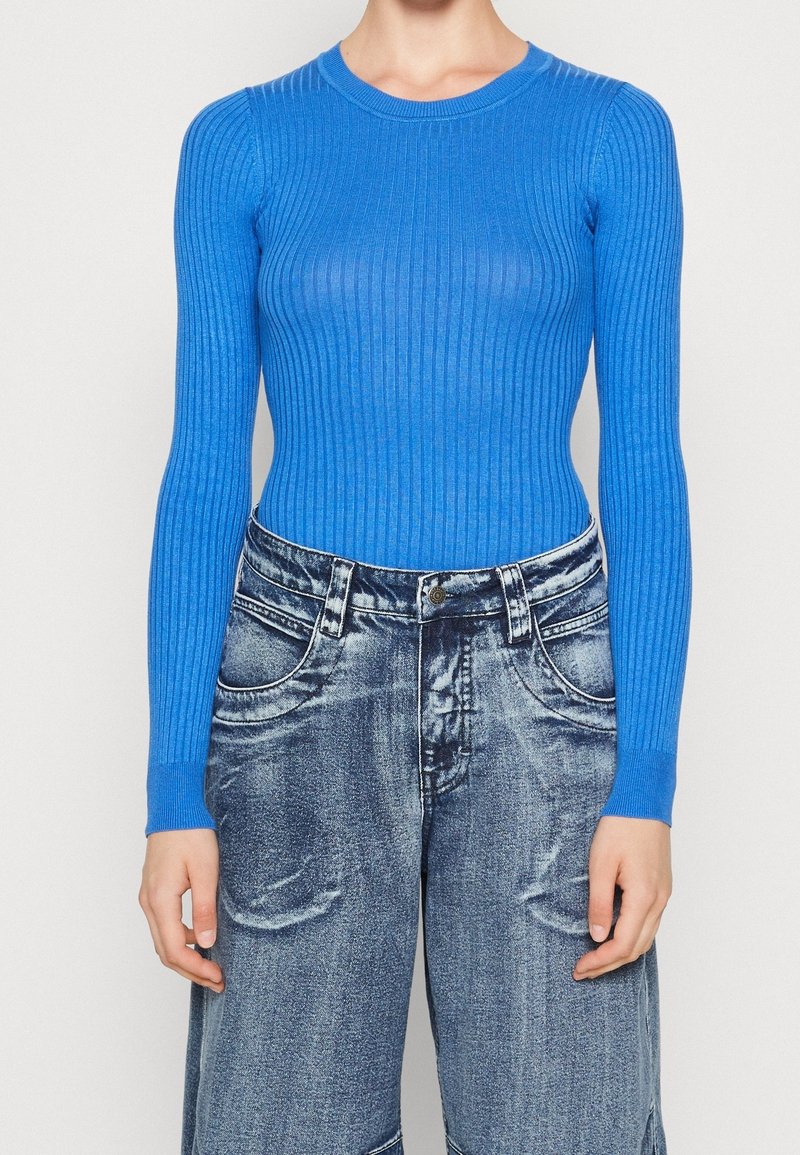 Personne portant un haut ajusté bleu vif en maille côtelée à manches longues, rentré dans un jean large bleu délavé taille haute. Mains détendues le long du corps.