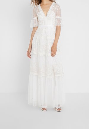 Valkoinen pitsikerroksinen maksimekko, jossa on läpikuultavat polkadot-kuviolliset kyynärpään mittaiset hihat, syvä V-aukkoinen pääntie ja kukkakirjailu, yhdistettynä valkoisiin sandaaleihin.