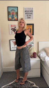 Juoda marškinėlių tipo palaidinė ir pilki cargo šortai su šoninėmis kišenėmis, šlepetės, stovintys ant raštuoto kilimo prieš šviesių spalvų sieną.