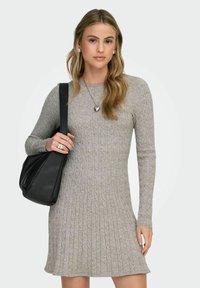 Femme aux longs cheveux blonds portant une robe en maille grise, un collier en forme de cœur, des boucles d'oreilles en perles, tenant un sac à main noir.