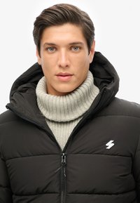 Sort dunjakke med hætte, med lynlås foran, syede paneler og et hvidt lynsymbol. Bæres over en lysegrå ribbet sweater.