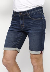 Homme portant un short en jean bleu foncé avec des revers retroussés et des coutures visibles, associé à une chemise blanche rentrée.
