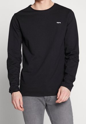 Mand iført en sort langærmet crewneck-trøje med lille hvidt "tigha"-logo på brystet, kombineret med lysegrå jeans.