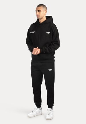 Schwarzer Hoodie mit Fronttasche und weißem Text, kombiniert mit passenden schwarzen Jogginghosen. Das Model trägt weiße Sneakers. Schlichte Gestaltung, Baumwollmischung.
