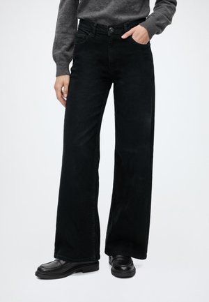 Persona que lleva pantalones negros de pierna ancha, una camiseta gris de manga larga y mocasines negros con una mano en el bolsillo sobre un fondo claro.