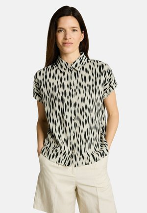 Kobieta z długimi ciemnymi włosami, ubrana w krótki rękaw, czarno-białą koszulę w kropki na guziki oraz jasnobeżowe krótkie spodenki z rękami w kieszeniach.