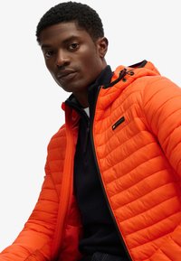 Chaqueta acolchada para hombre en un vibrante color naranja, con textura acolchada, capucha, cierre con cremallera y un pequeño detalle del logo en el pecho.