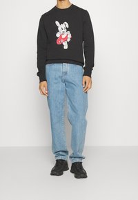 Zwarte sweatshirts met een cartoonkonijntje dat een rood voorwerp vasthoudt, gecombineerd met lichtblauwe spijkerbroeken en zwarte sneakers.