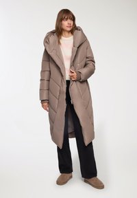 Długa brązowa kurtka puchowa z kapturem, wypełniona, z przekątnym przeszyciem i zamkiem z przodu. Noszona na czarnych spodniach i brązowym obuwiu.