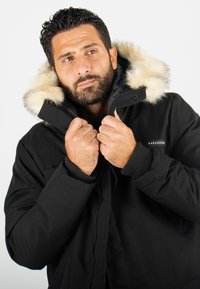 Veste d'hiver noire avec une capuche doublée de fourrure, fermeture éclair et poignets côtelés. Présente un logo sur la poitrine et une texture lisse et matte.