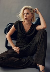 Sort og guldfarvet metallisk jumpsuit med draperet halsudskæring, brede ben og indsnævret talje. Modellen læner sig imod en sort stol og fremviser stoffets glans.