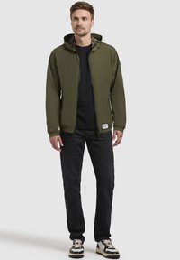 Olivegrüne, leichte Jacke mit Reißverschluss, verstellbarem Kapuzenkragen und elastischen Bündchen, getragen über einem schwarzen Hemd und kombiniert mit schwarzen Jeans.