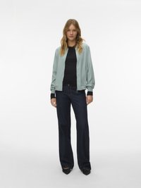 Γυναίκα με κυματιστά ξανθά μαλλιά που φοράει ανοιχτοπράσινο bomber μπουφάν, μαύρο μακρυμάνικο τοπ, σκούρα μπλε τζιν φαρδιάς γραμμής και μαύρα μυτερά παπούτσια.