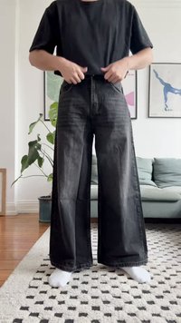 Jeans de ganga pretos de perna larga com uma cintura alta, apresentando detalhes desbotados e uma bainha desfiada. Usados com uma t-shirt preta e meias brancas.
