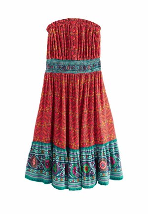 Femme en robe bustier rouge à motifs avec bordure florale bleu sarcelle, debout tenant un sac cabas gris tissé près d'une porte en bois abîmée.