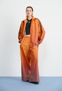 Oransje treningsjakke med svarte striper, sammen med matchende bukser med ombre-effekt. Svart croppet topp under, svart fottøy.