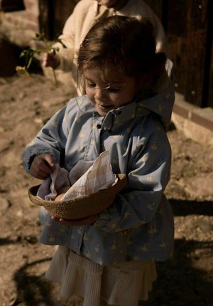 Jonge kind in een blauwe jas houdt een mand met een doek vast en plaatst voorzichtig een voorwerp erin of haalt dit eruit, staand buiten in het zonlicht.
