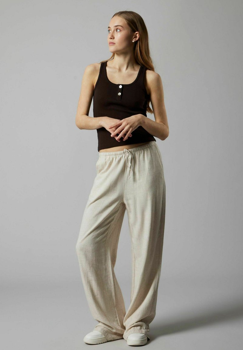 Zwarte geribbelde tanktop met drie knopen, gecombineerd met lichtgewicht crèmekleurige broek met trekkoord en witte sneakers.