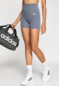 Pantaloni scurți atletici gri cu talie înaltă și fit strâns, cu logo-ul alb Adidas. Modelul ține o geantă de sală neagră cu inscripționare albă Adidas.