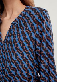 Blaue und schwarze gemusterte Bluse mit wellenförmigem Design und braunen Akzenten, Knopfverschluss und gefälteltem Ausschnitt. Weicher Stoff.