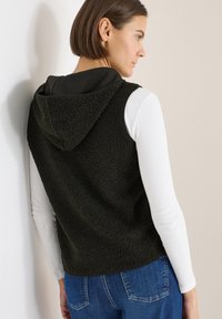 Dunkelgrüne Fleeceweste mit Kapuze und Struktur, kombiniert mit einem weißen Langarmshirt. Verfügt über eine lockere Passform und einen geformten Saum.