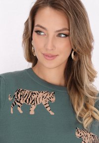 Pulover verde cu un model de tigru tricotat în nuanță bej. Are un guler rotund și margini cu nervuri. Textură netedă, design ajustat.
