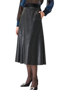 Femme portant une jupe midi en cuir noire en A, des collants noirs transparents, des talons en satin noirs avec des boucles en cristal et un chemisier transparent avec des manches bleues.