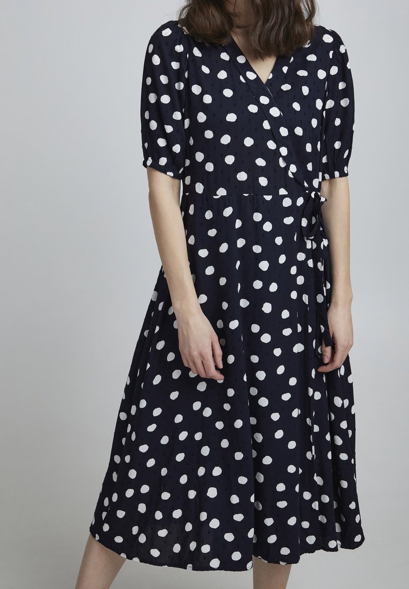 Femme portant une robe portefeuille bleu marine avec des manches courtes bouffantes et de grands pois blancs, debout devant un fond gris uni.