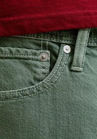 Gros plan d'une poche de jean en denim vert avec rivets argentés et passants de ceinture, associé à un ourlet de chemise en tissu côtelé rouge au-dessus.