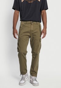 Khaki spodnie cargo z wieloma bocznymi kieszeniami, dopasowanym pasem i zakończeniami nogawek w mankiety, zestawione z czarnym t-shirtem z grafiką i białymi trampkami za kostkę.