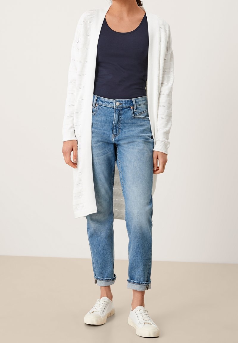 White open-knit cardigan over a navy tank top, paired with light-wash, rolled-cuff jeans and white sneakers. Simple, casual look.